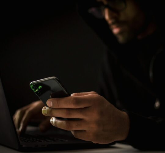 Crop anonymous ethnic male cyber spy with cellphone and netbook hacking system in evening