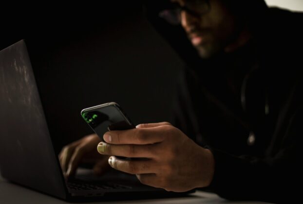 Crop anonymous ethnic male cyber spy with cellphone and netbook hacking system in evening
